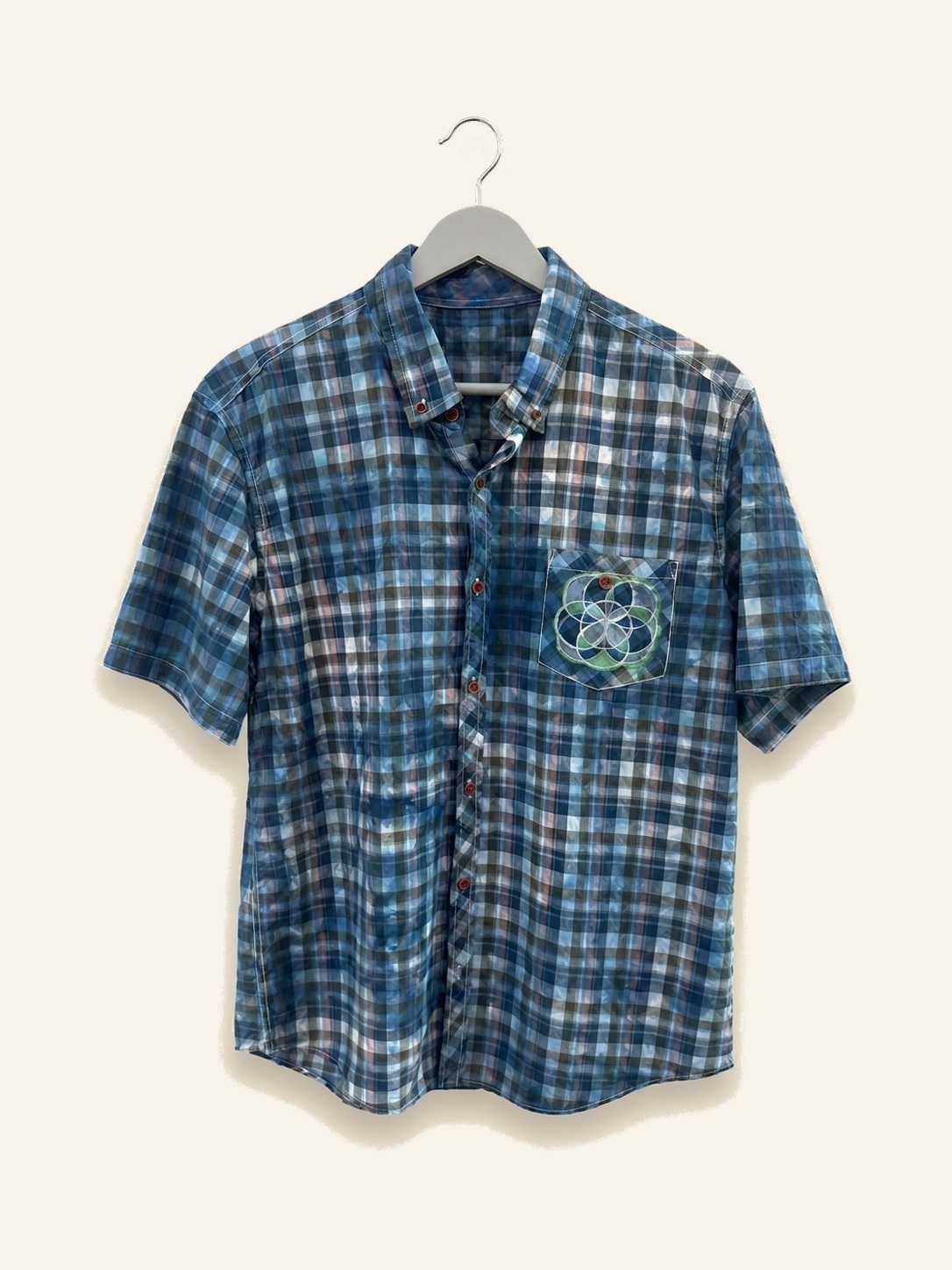 Hand ice-dyed cotton blue plaid shirt with a green sacred geometry emblem on the pocket, on a pale grey hanger